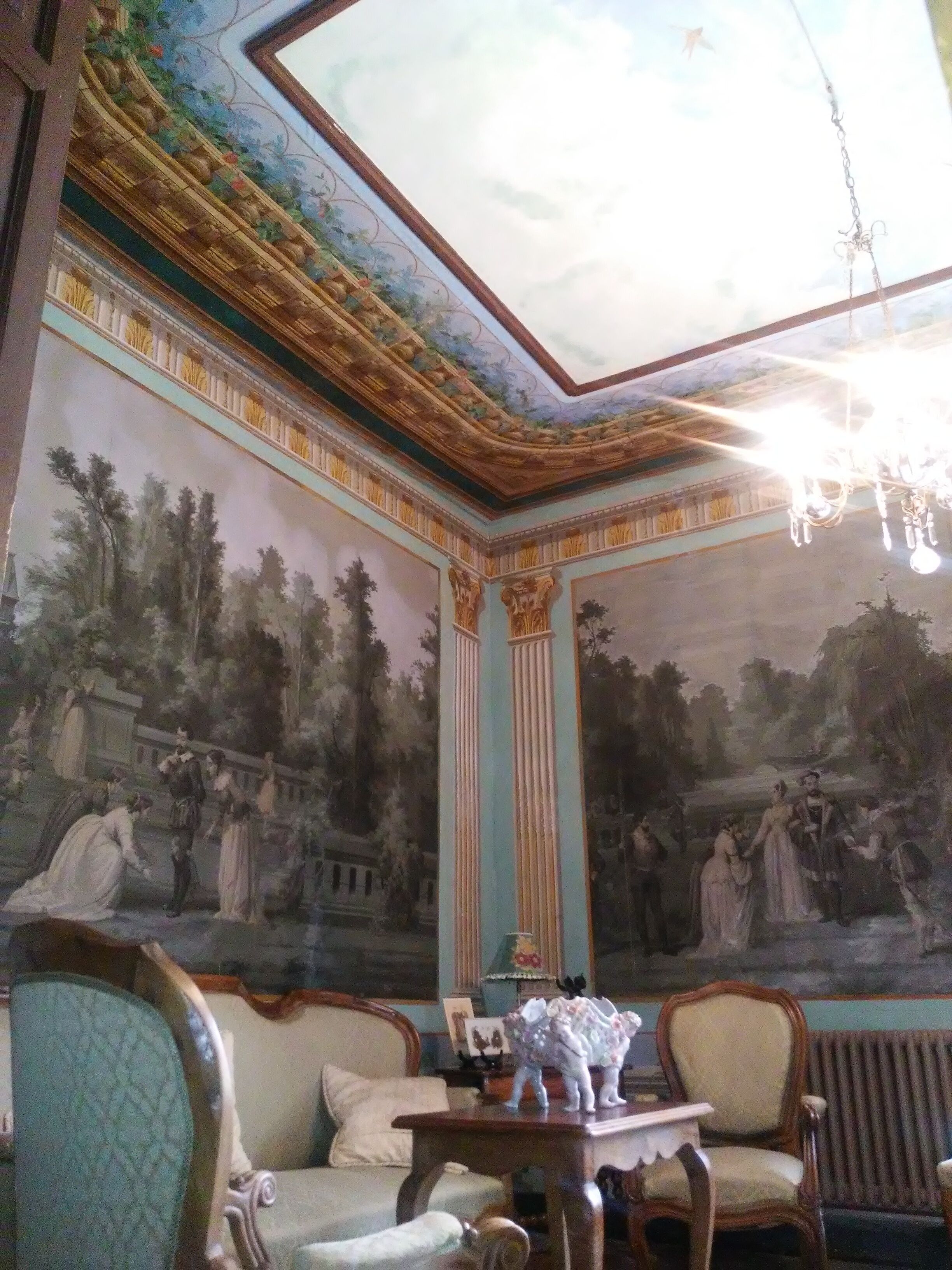 Una habitación con un mural, techo ornamentado y muebles clásicos.