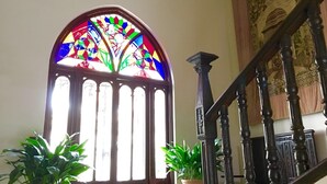 Una ventana de vitral con diseños geométricos, una escalera de madera con barandales ornamentales y dos plantas en macetas.