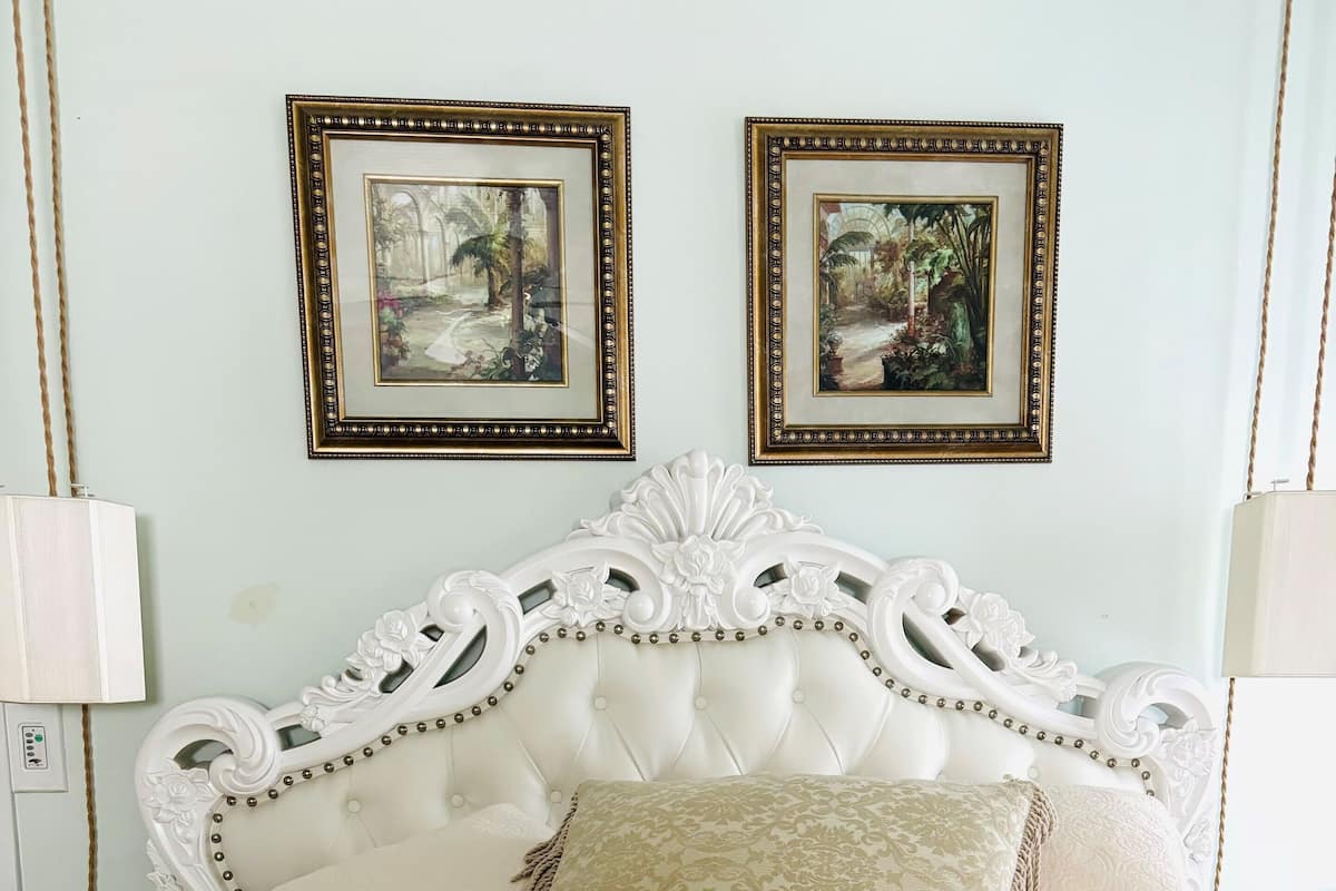 A bed with a white headboard, two framed pictures on the wall, and a bedside lamp.