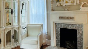 Una habitación con un sillón blanco, una chimenea blanca y un mueble grande con puertas de vidrio.