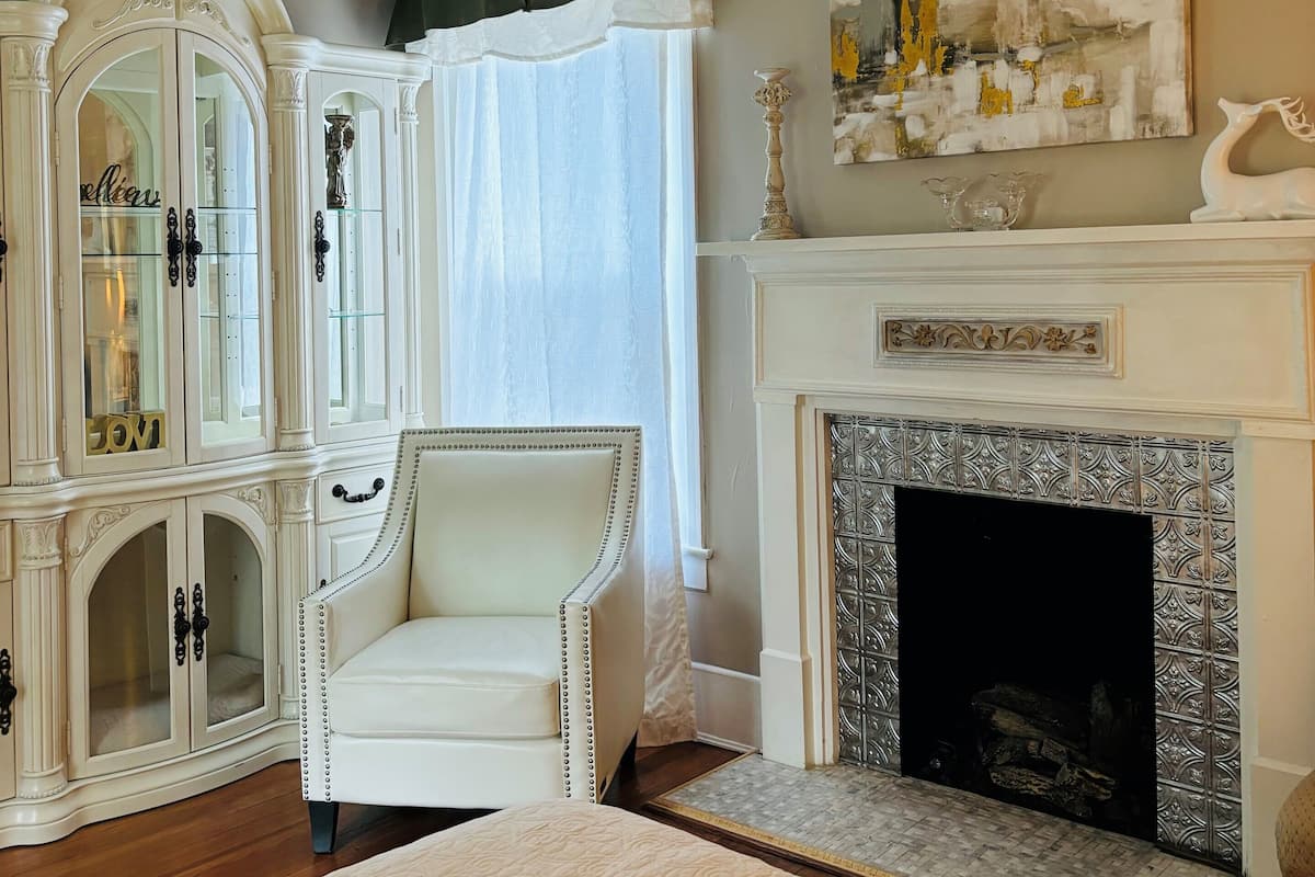 A room with a white armchair, a white fireplace, and a large cabinet with glass doors.