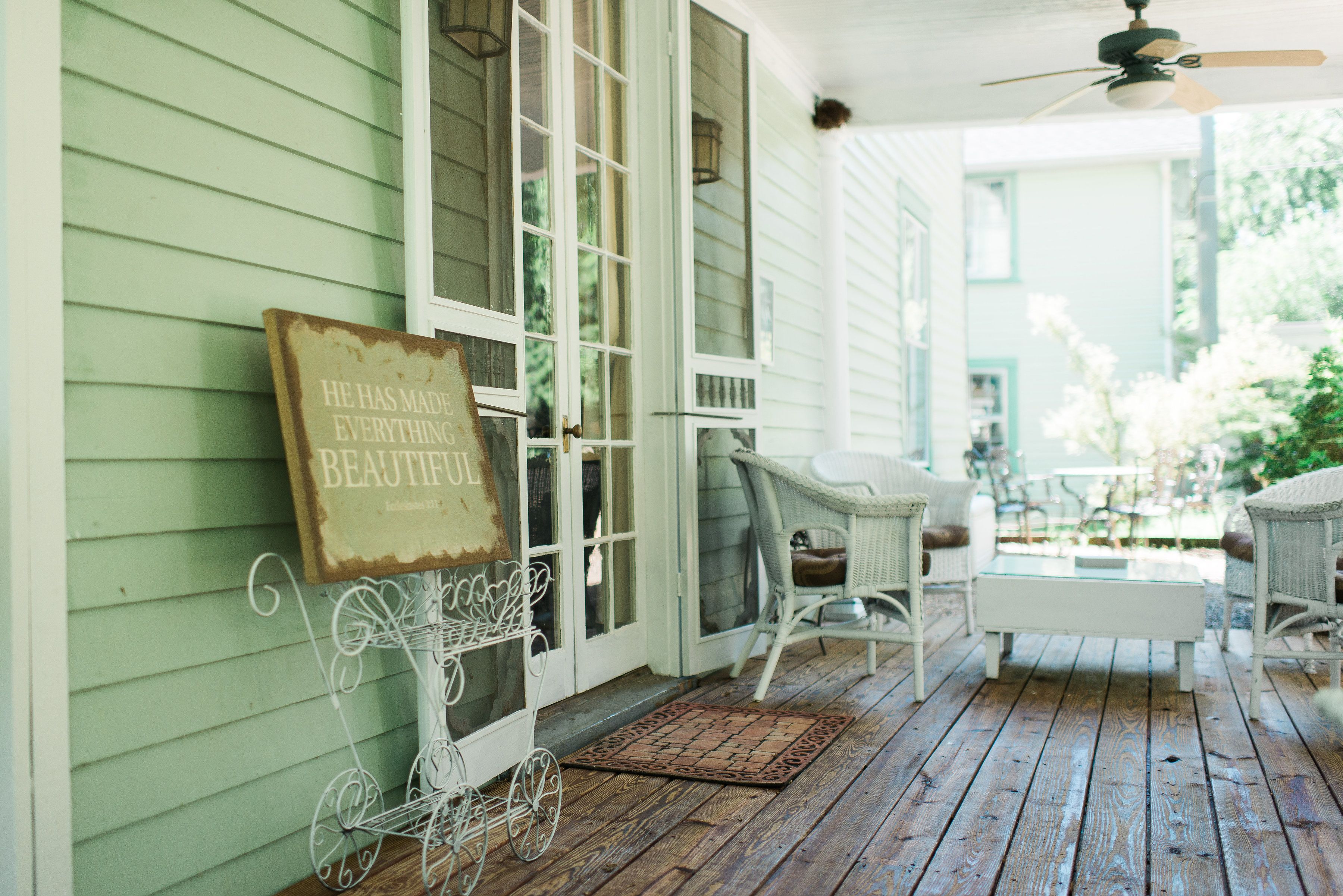 A porch with wicker furniture, a white bench, and a sign that reads "He has made everything beautiful."