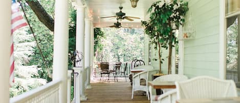 A covered porch with wooden flooring, white wicker furniture, ceiling fans, and a view of greenery.