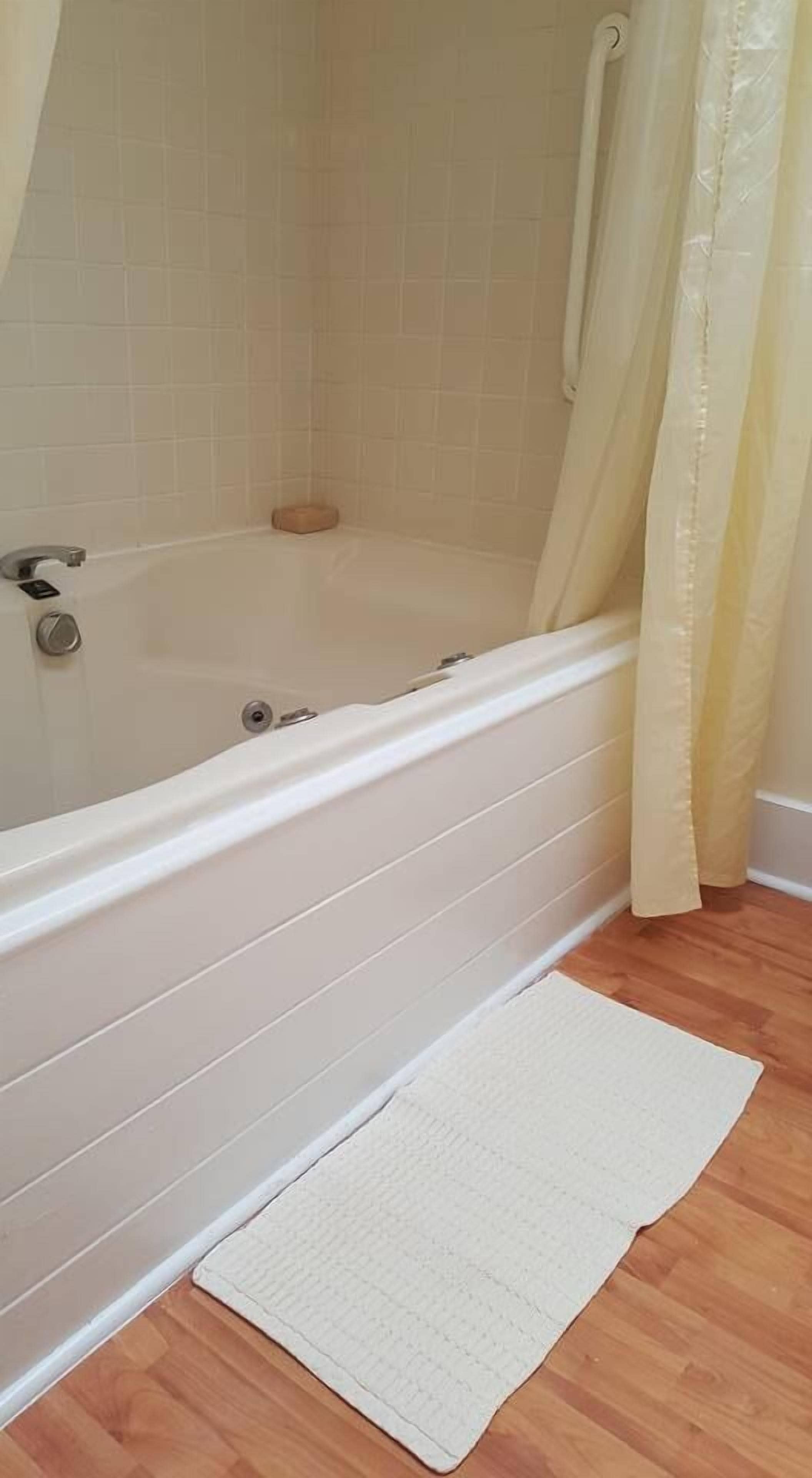 Un baño con una bañera blanca, una cortina de ducha y un tapete de baño blanco sobre un suelo de madera.