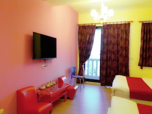 Una habitación con televisión montada en la pared, un sofá rojo, una mesita con adornos, un balcón con cortinas estampadas y una cama con una colcha roja.