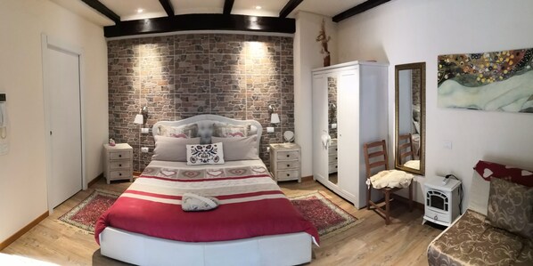 A bedroom with a stone wall, a bed with a red and white bedspread, a white wardrobe, and a small heater on the floor.