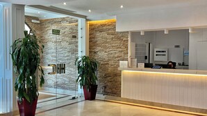 A reception area with a stone wall, a glass door marked 'SALIDA DE EMERGENCIA,' and a reception desk with a person seated behind it. - Hotel Castilla Torrijos (Torrijos)