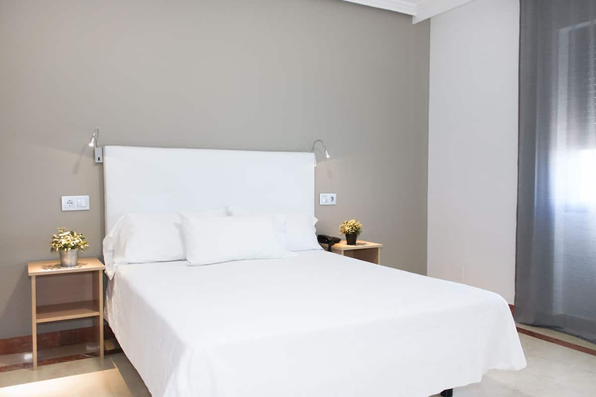 A neatly made bed with white linens in a hotel room. There are two bedside tables, one with a vase of flowers and the other with a phone. The room has a simple, clean design with neutral walls.