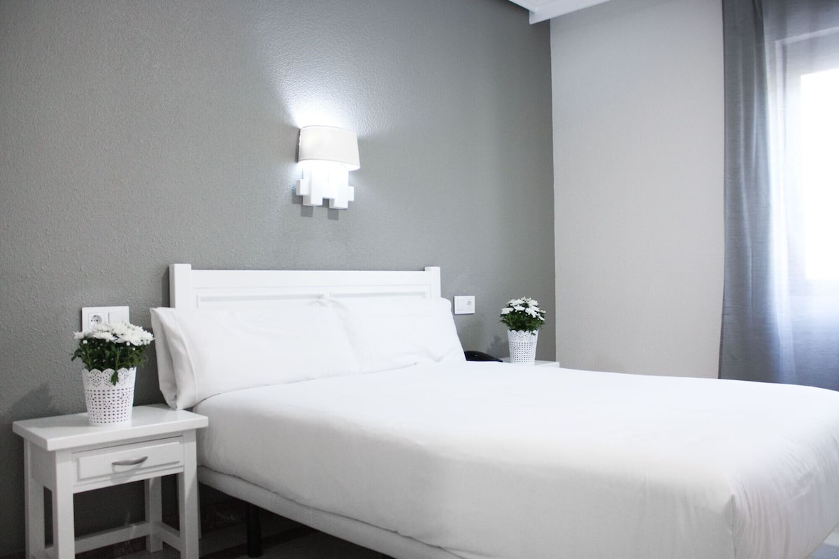 A neatly made bed in a room with grey walls, a white nightstand with a lamp, and a small white vase with flowers.