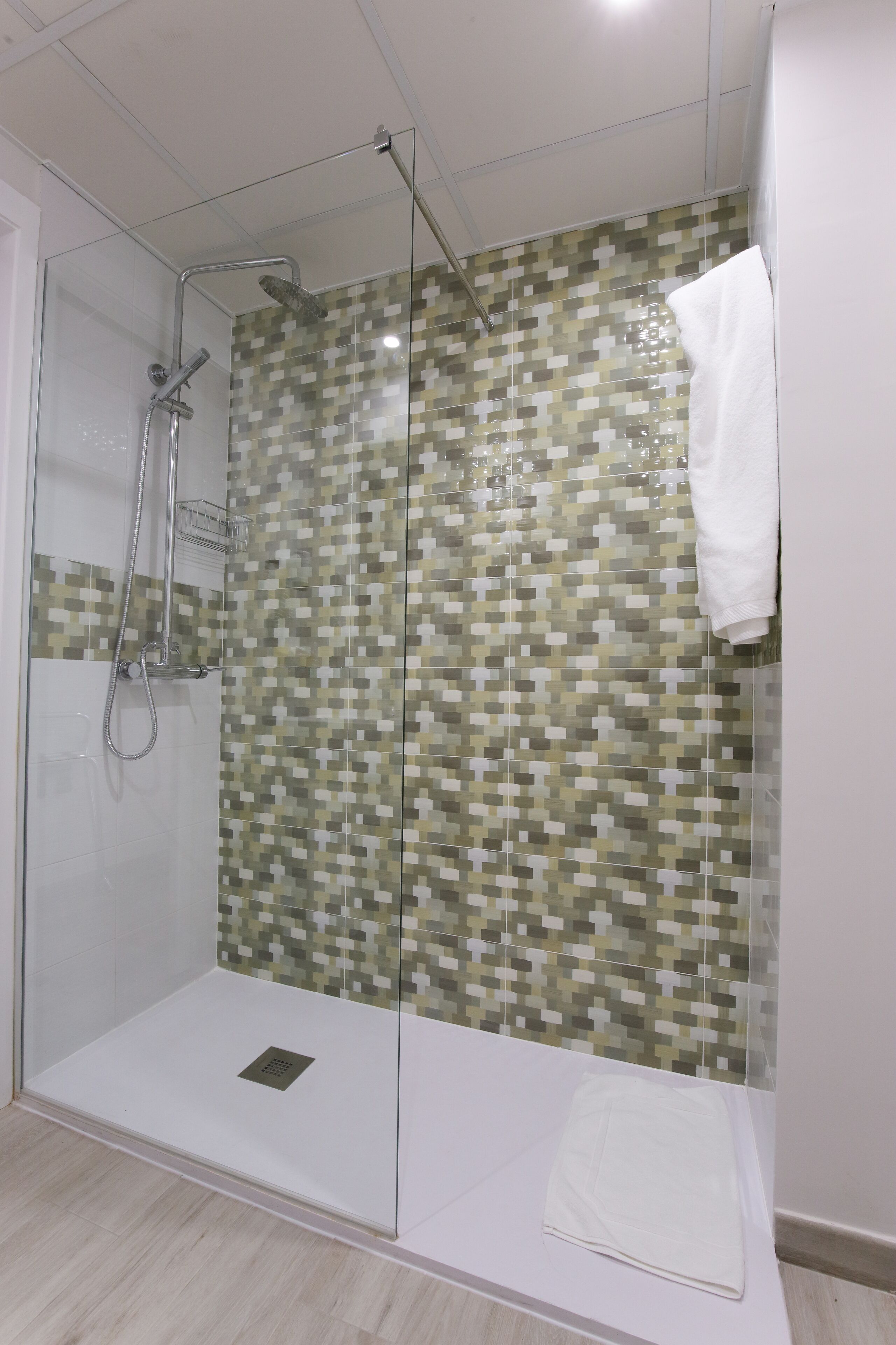 A glass-enclosed shower with a tiled mosaic wall, a white towel, and a showerhead.
