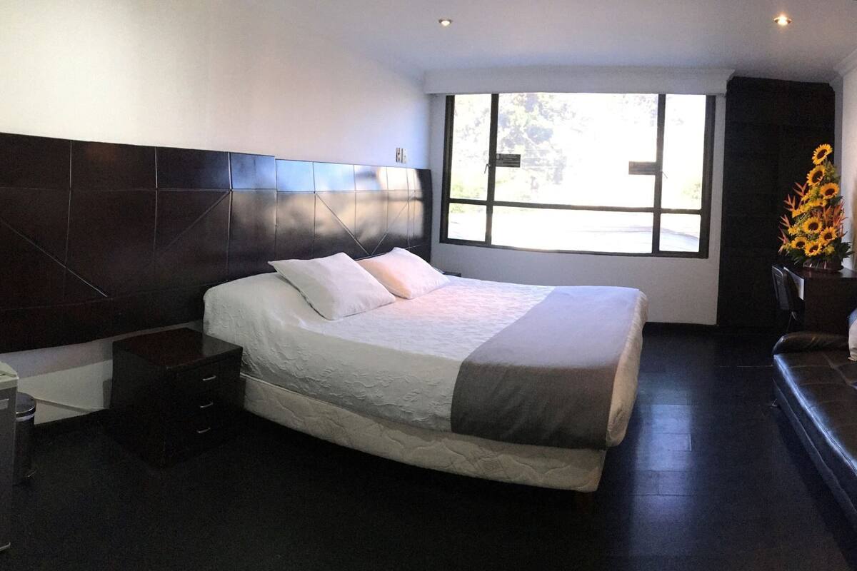 A modern hotel room with a large bed, a nightstand, a dresser, and a window with a view.
