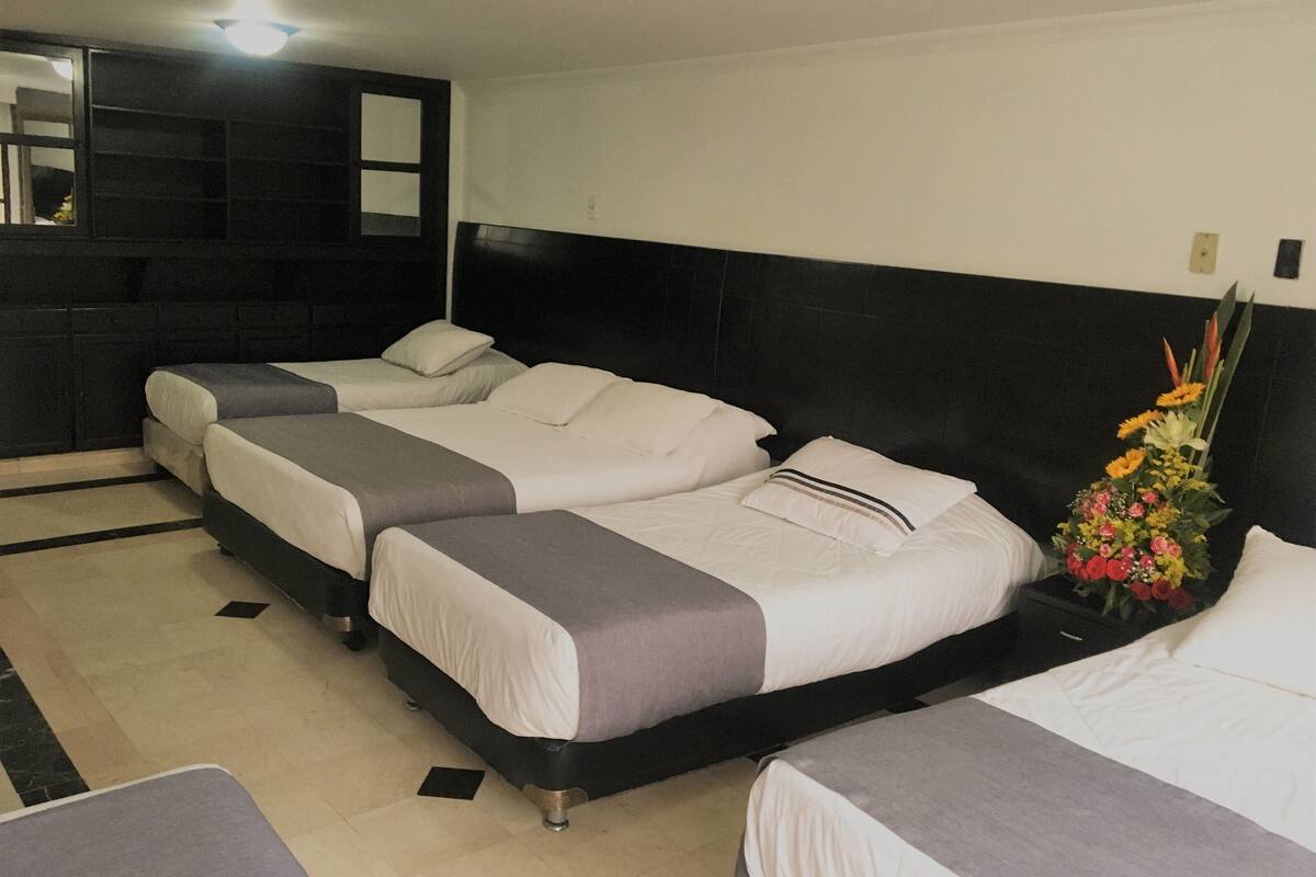 A hotel room with three beds arranged in a row, a black headboard, a small bedside table with a floral arrangement, and a mirror on the wall.