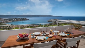 A dining table set with plates, cutlery, and a pitcher of juice, overlooking a coastal landscape with buildings and the sea. - Sounio Villa (Lavreotiki)