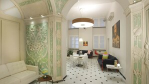 Ein geräumiges Wohnzimmer mit einem weißen Sofa, einem runden Couchtisch und einem Essbereich mit Tisch und Stühlen. Der Raum zeichnet sich durch aufwändige, grüne und weiße Wandverzierungen sowie eine hohe Decke mit einer großen, runden Leuchte aus.