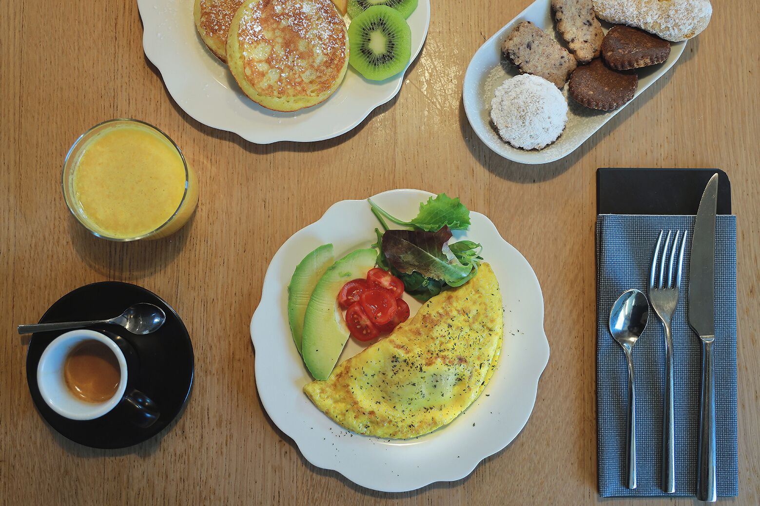 Un desayuno con tortitas, kiwi, galletas, una tortilla de aguacate y tomate, un vaso de zumo de naranja y una taza de café.