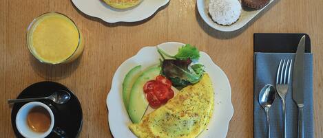 Un desayuno con tortitas, kiwi, galletas, una tortilla de aguacate y tomate, un vaso de zumo de naranja y una taza de café.