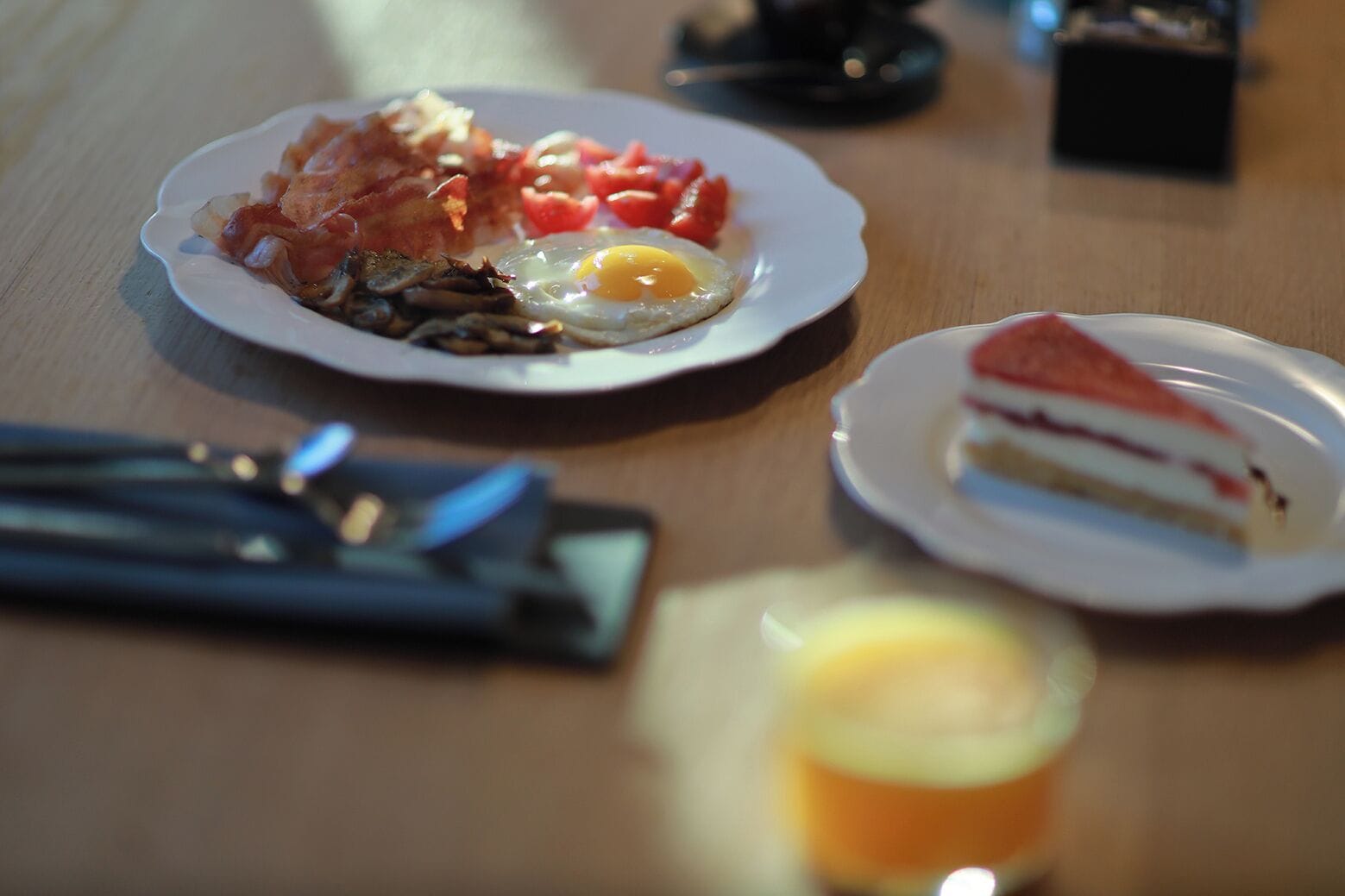A breakfast plate with bacon, eggs, and tomatoes, a slice of cake, and a glass of orange juice on a wooden table.