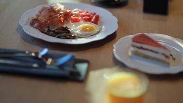 A breakfast plate with bacon, eggs, and tomatoes, a slice of cake, and a glass of orange juice on a wooden table.
