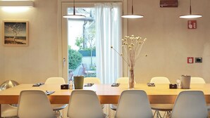Ein Essbereich mit einem langen Tisch, Stühlen, Pendelleuchten, einer Vase mit getrockneten Blumen und einem Bild an der Wand.