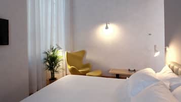 A hotel room with a bed, a yellow armchair, a small table with a lamp, and a potted plant.