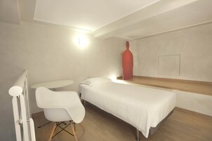 Een minimalistische slaapkamer met een enkel bed, een witte stoel en een rode sculptuur.