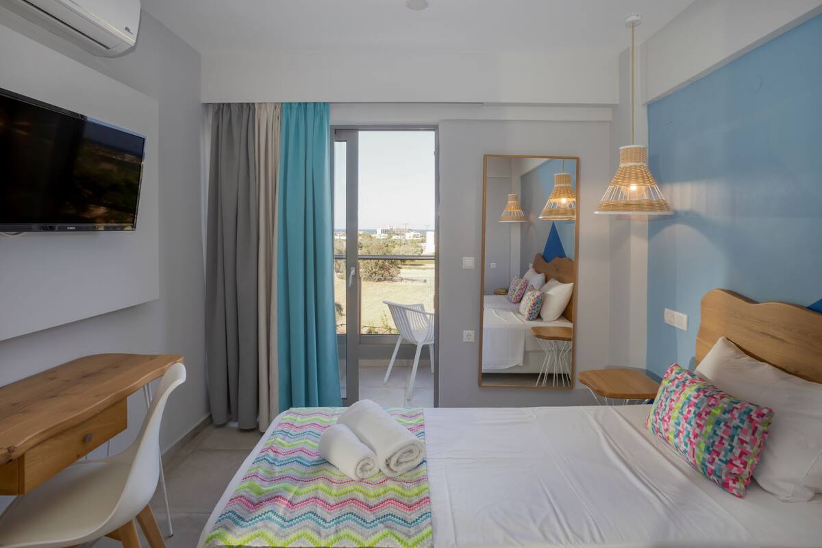 A hotel room with a bed, a desk, a chair, a TV, a balcony with a view, and a mirror.