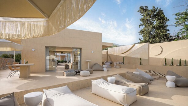 Un área lounge exterior moderna con sofás y cojines color beige, una mesa de piedra y una entrada con puerta de vidrio.
