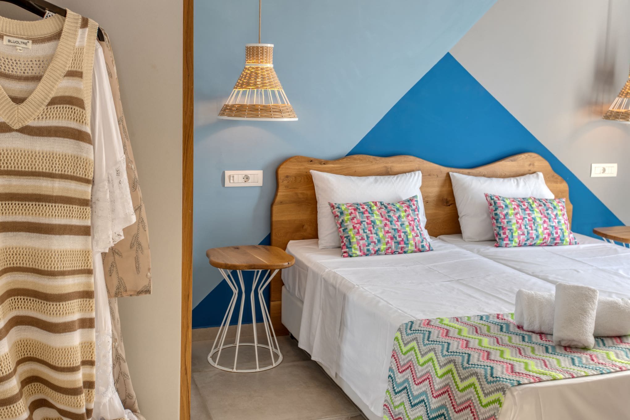 A hotel room with a bed, two pillows, a bedside table, a hanging light, and a wall with a geometric design.