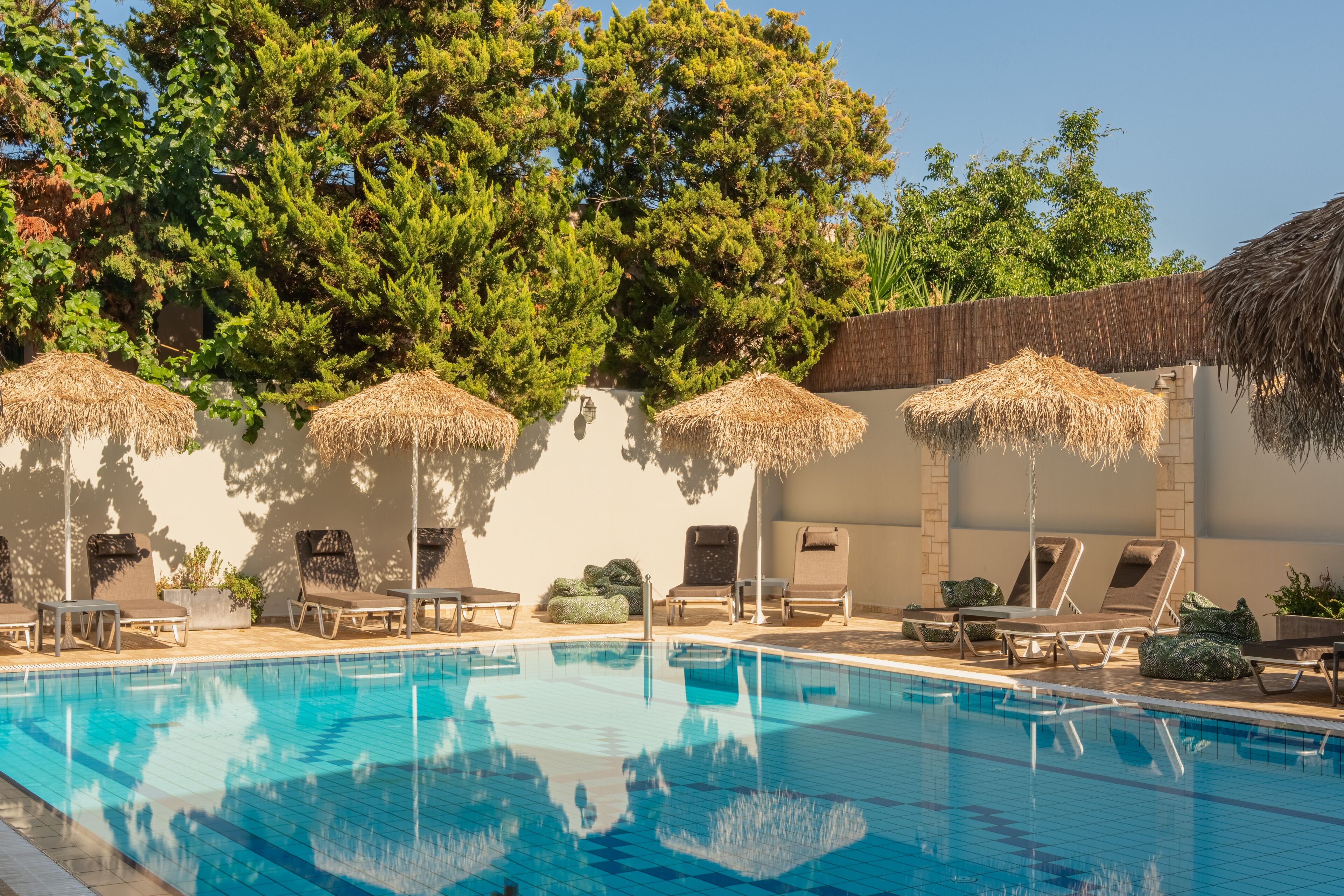 A swimming pool with lounge chairs and thatched umbrellas, surrounded by trees and a stone wall.