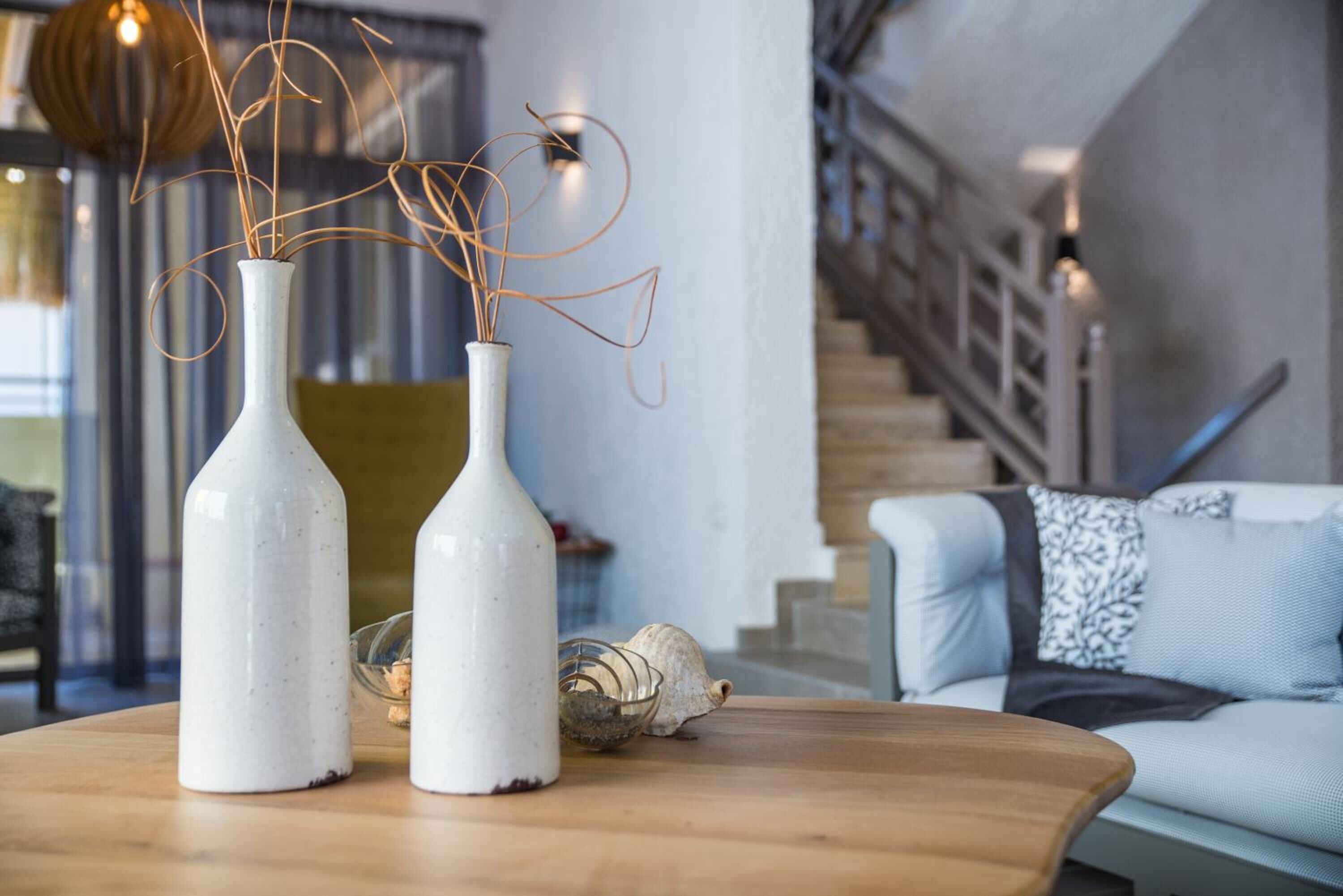 Two white ceramic vases with twisted branches on a wooden table, a shell, and a staircase in the background.