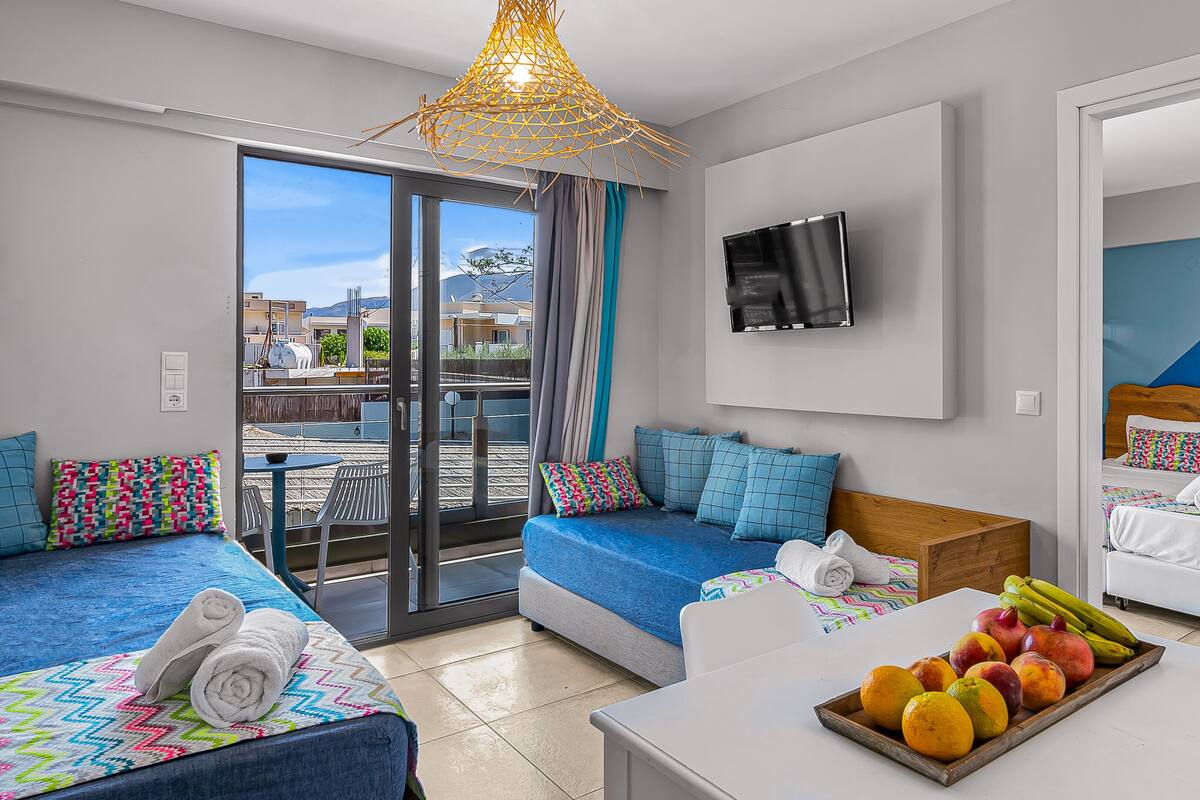 A modern hotel room with two beds, a sofa, a dining table, and a fruit bowl.