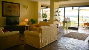 Un vaste salon comprenant un grand canapé, un téléviseur et un tableau accroché au mur. Une porte vitrée donne accès à un espace extérieur d’où l’on peut admirer le paysage.