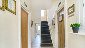 Un pasillo con una puerta de madera, un letrero de recepción, un cartel de 'No fumar', una planta en maceta y una escalera.