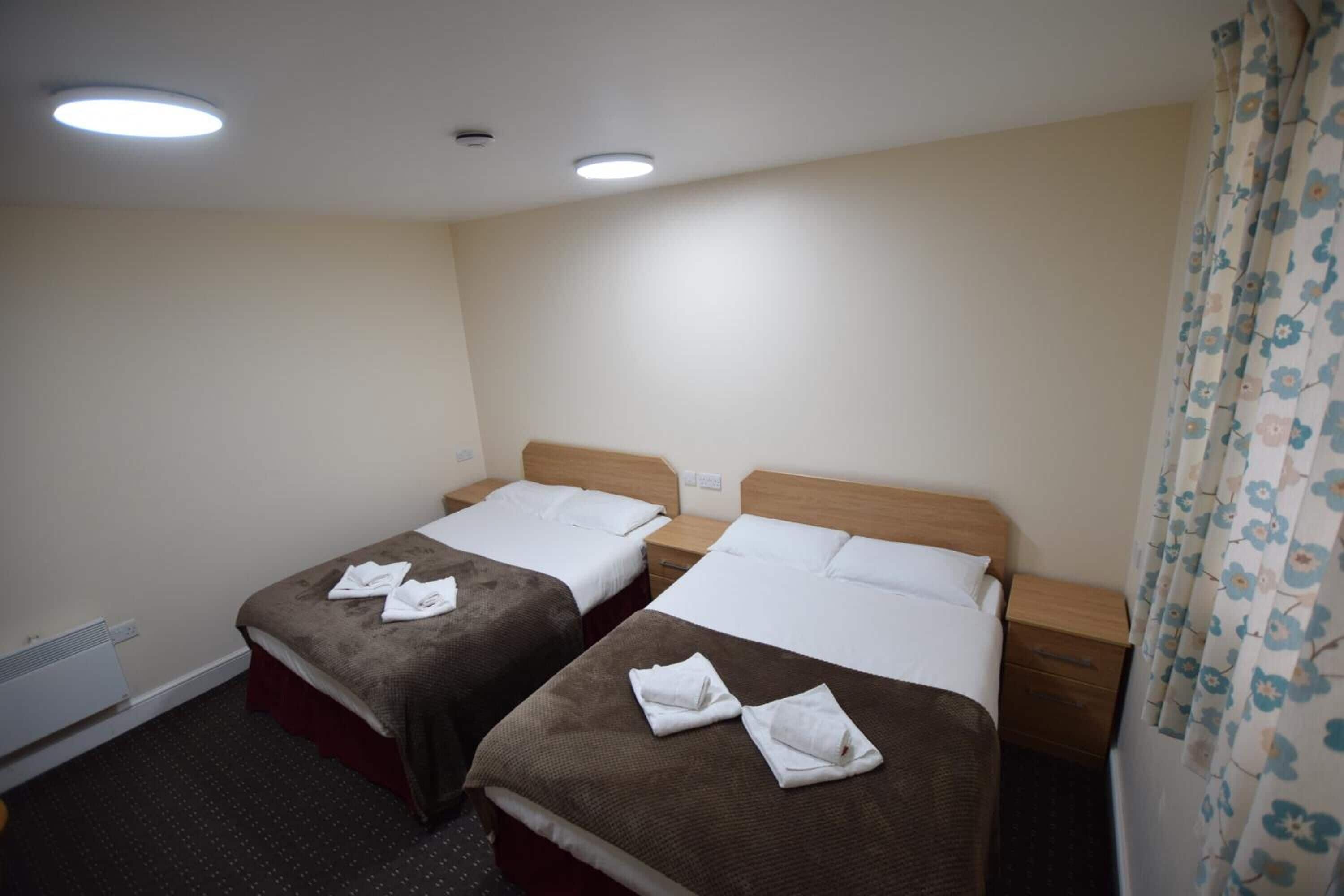 A hotel room with two beds, each with a brown bedspread and white linens, and a wooden headboard. There is a small bedside table with a lamp on each side of the beds.