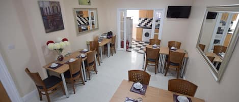 A dining area with multiple tables and chairs, a kitchen visible through an open door, and a clock on the wall.