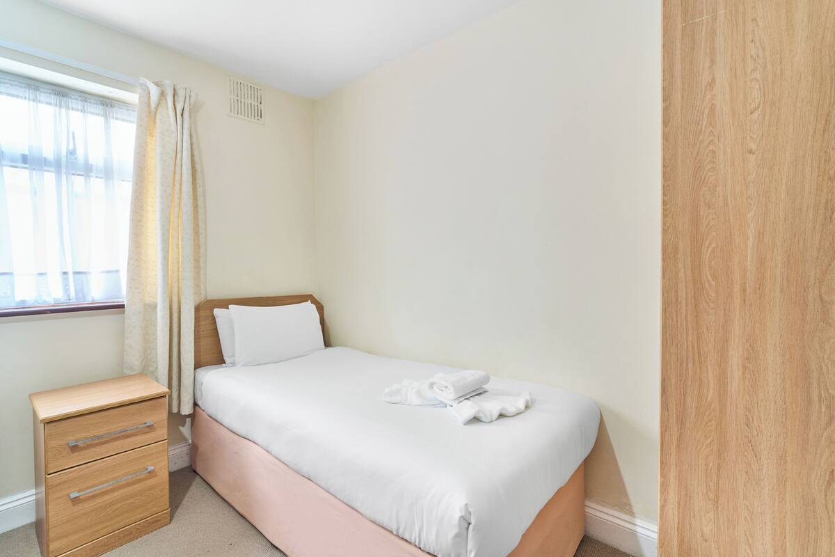 A neatly made bed with white linens, a wooden nightstand with drawers, and a window with sheer curtains.