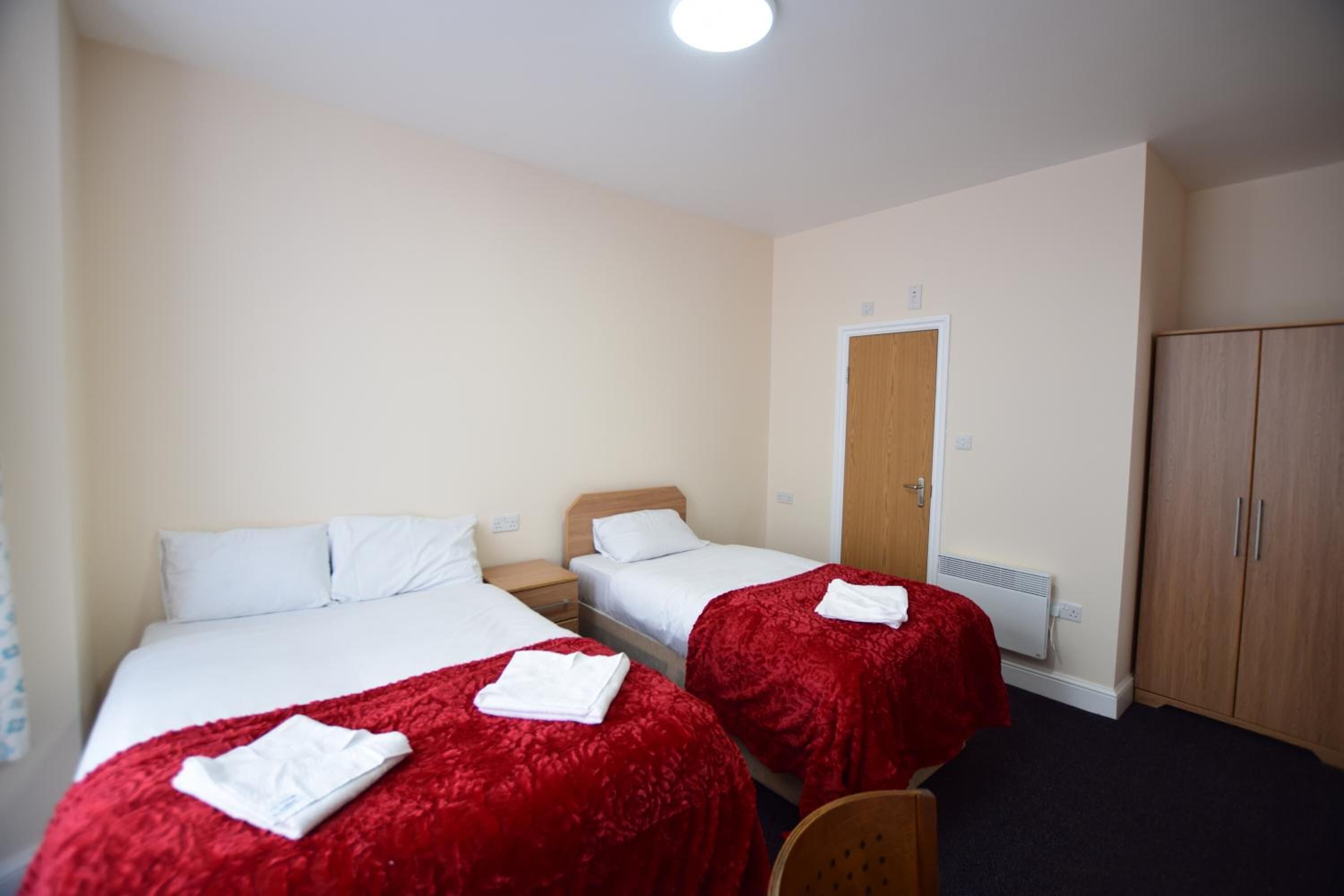 A hotel room with two beds, each with a red blanket and white pillows, a wooden wardrobe, and a door leading to another room.