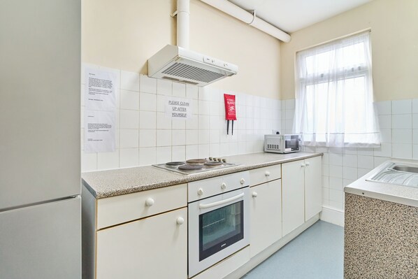 A kitchen with white cabinets, a stove, a microwave, and a window with sheer curtains. - Woodlands Lodge Ilford (Ilford)
