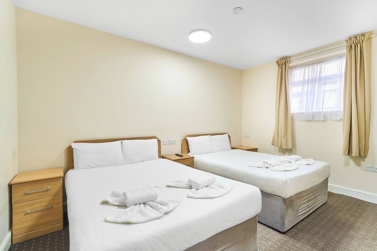 A hotel room with two beds, a wooden nightstand, a drawer unit, and a window with curtains.