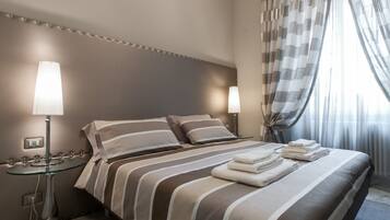 A neatly made bed with striped bedding and folded towels, a bedside table with a lamp, and a view of the room through sheer curtains.