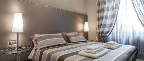 A neatly made bed with striped bedding and folded towels, a bedside table with a lamp, and a view of the room through sheer curtains.