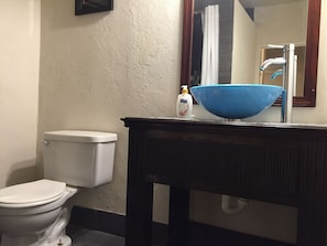 Un baño con inodoro blanco, lavamanos azul sobre un mueble de madera oscura y una botella de jabón.