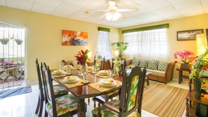 A dining area with a table set for four, surrounded by chairs with floral upholstery. There is a ceiling fan above the table, and a sofa with a patterned cushion is visible in the background. - BK Villas (Maxwell)