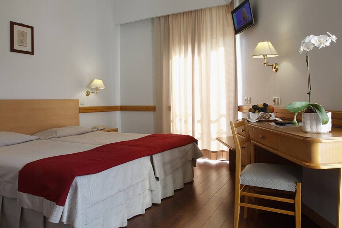 A hotel room with two beds, a desk with a chair, a television, and a cross on the wall.
