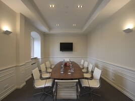Un salón de conferencias con una mesa larga, sillas blancas, un televisor de pantalla plana y luces empotradas en el techo.