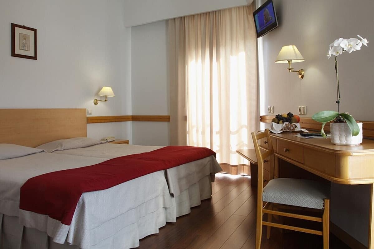 A hotel room with a bed, a desk, a chair, a television, and a cross on the wall.