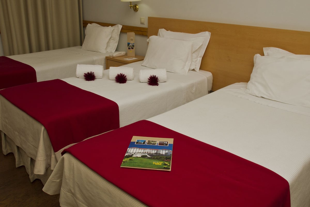 A hotel room with two beds, each with white bedding and red throw pillows. There is a nightstand with a lamp and a phone, and a brochure on the bed.