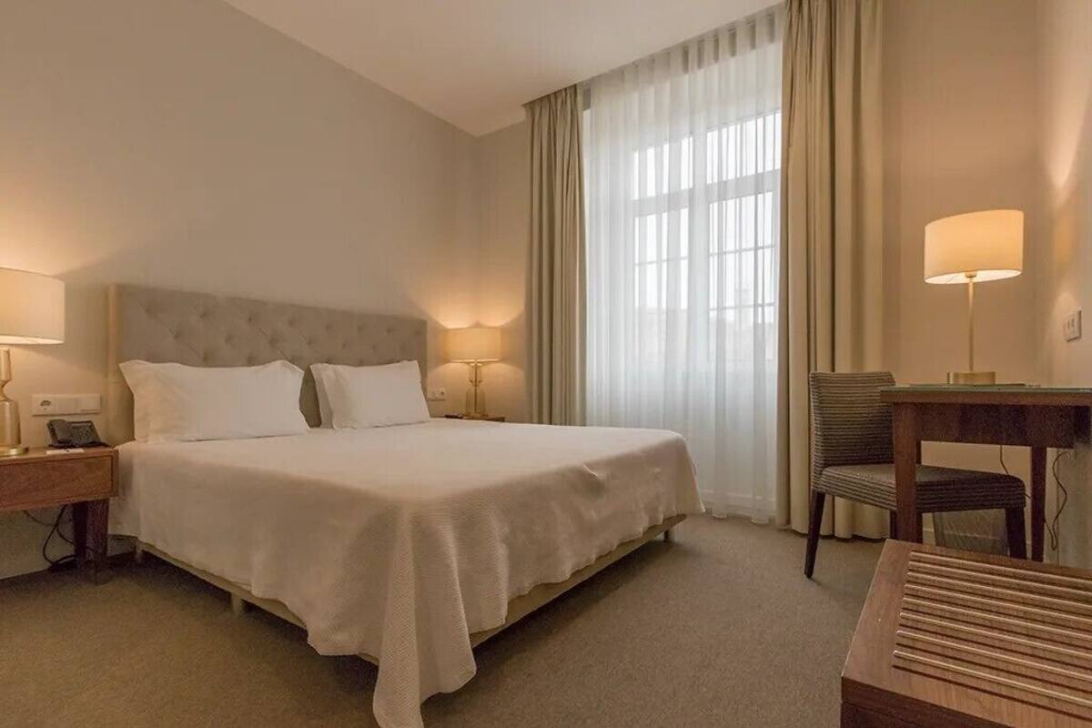 A hotel room with a large bed, two bedside tables with lamps, a desk with a chair, and a large window with curtains.