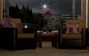 Una habitación con dos sillones, una mesita con un jarrón de flores y un ventanal con vistas a edificios y árboles.