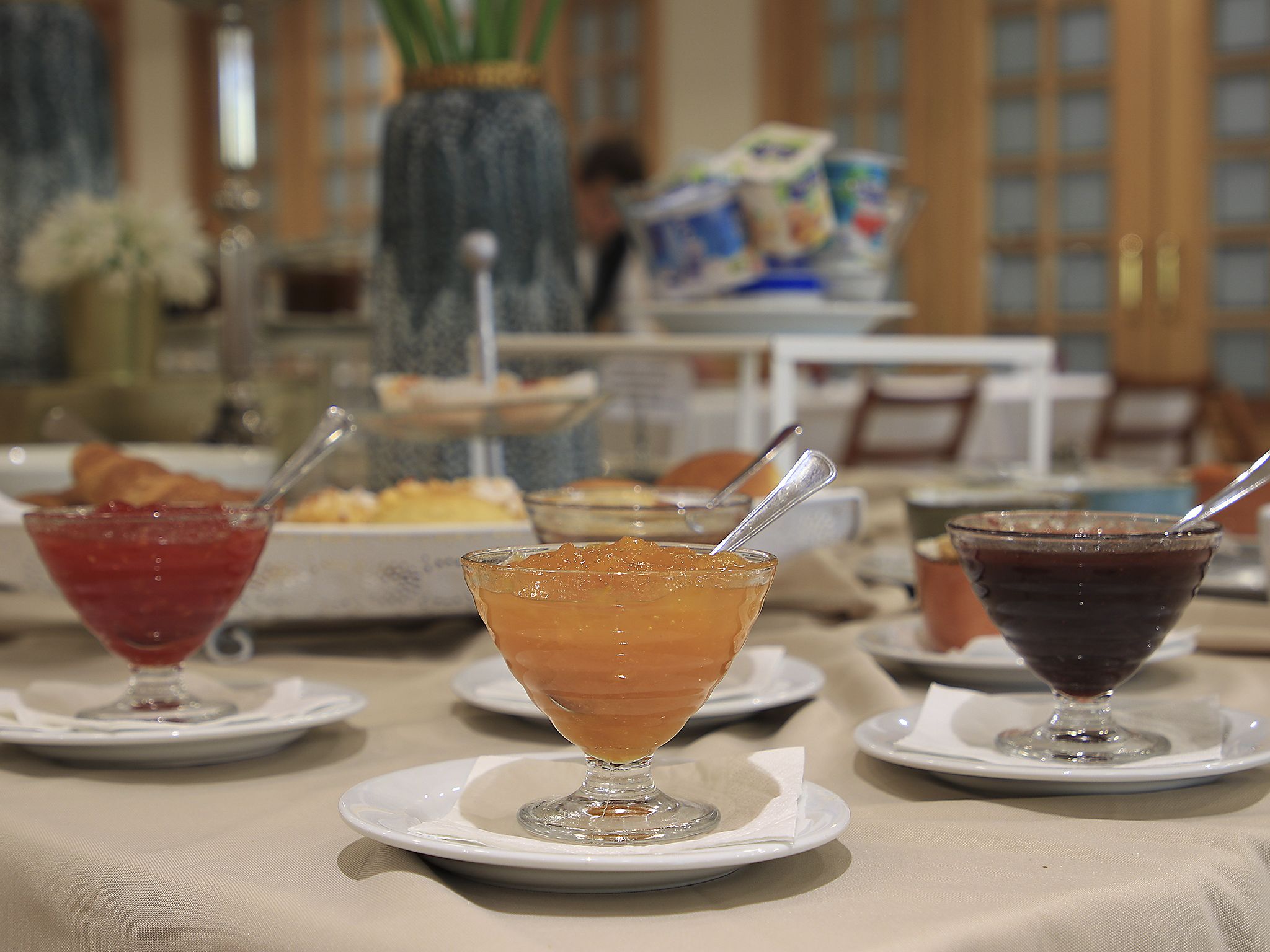 Ein Tisch mit drei Dessertgläsern, die verschiedene Getränke enthalten, eine Vase mit Blumen und ein Speisebereich im Hintergrund.